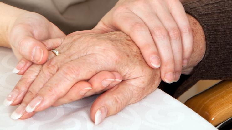 Zwei Hände, eine ältere und eine jüngere, liegen auf einem Tisch, wobei die jüngere Hand die ältere sanft umschließt.